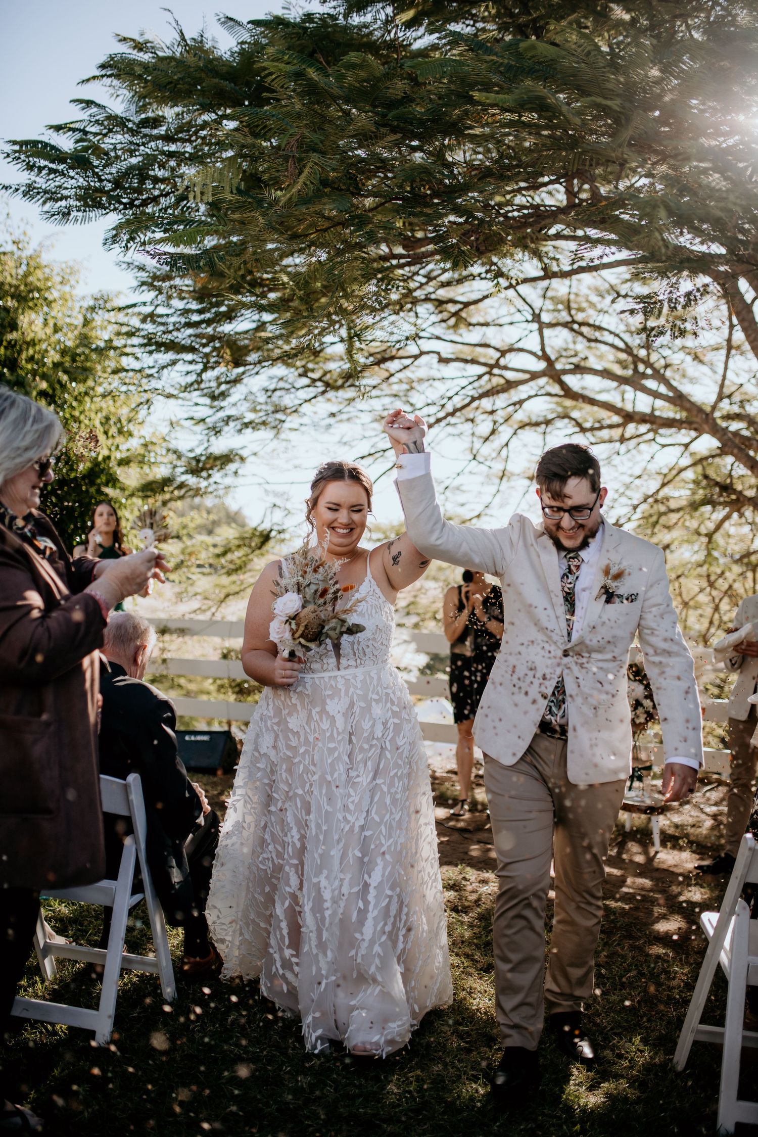 Newlyweds celebrate with raised hands while confetti falls during their outdoor ceremony.