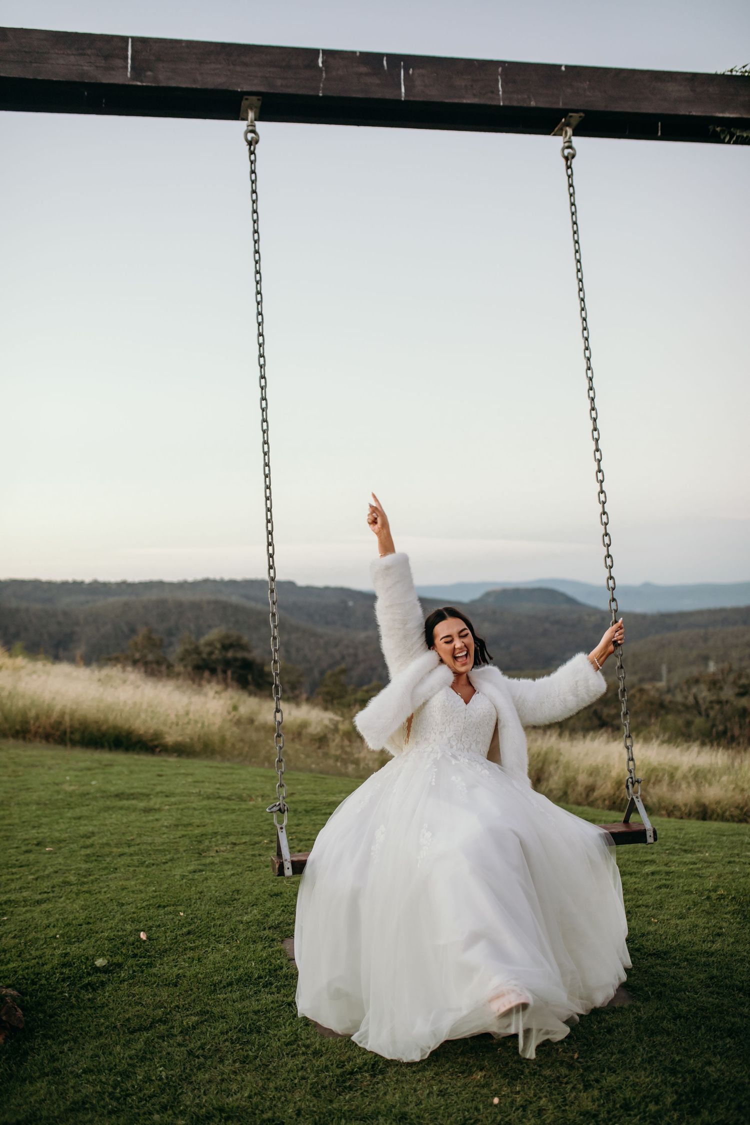 Joyful bride in white dress swings outdoors with mountain landscape in background.