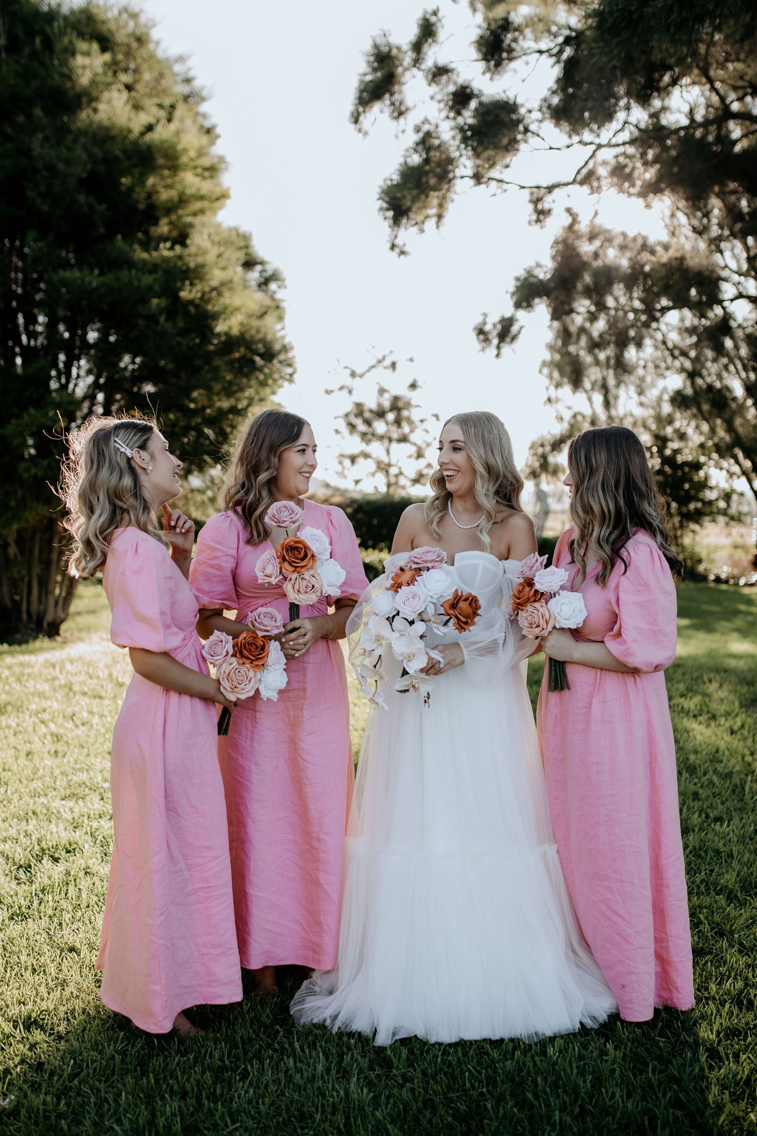 Bridesmaids in pink dresses gather around a bride in white, holding bouquets in an outdoor setting.