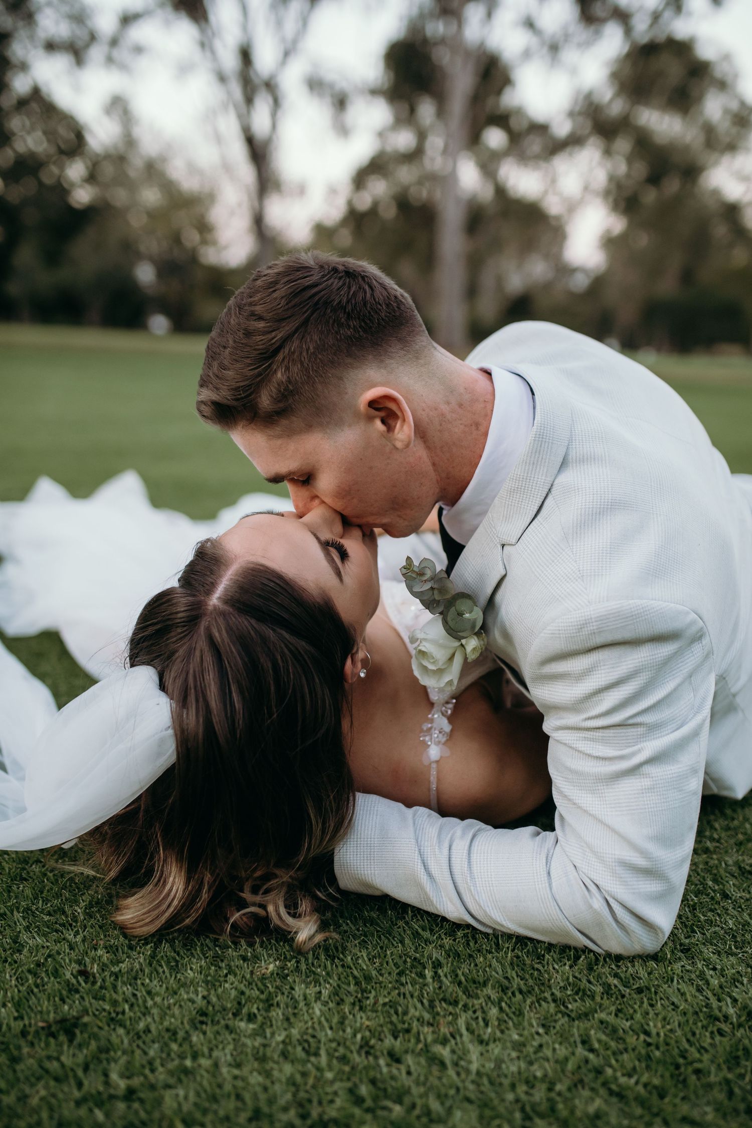 A romantic wedding couple lying together on green grass in a park setting.