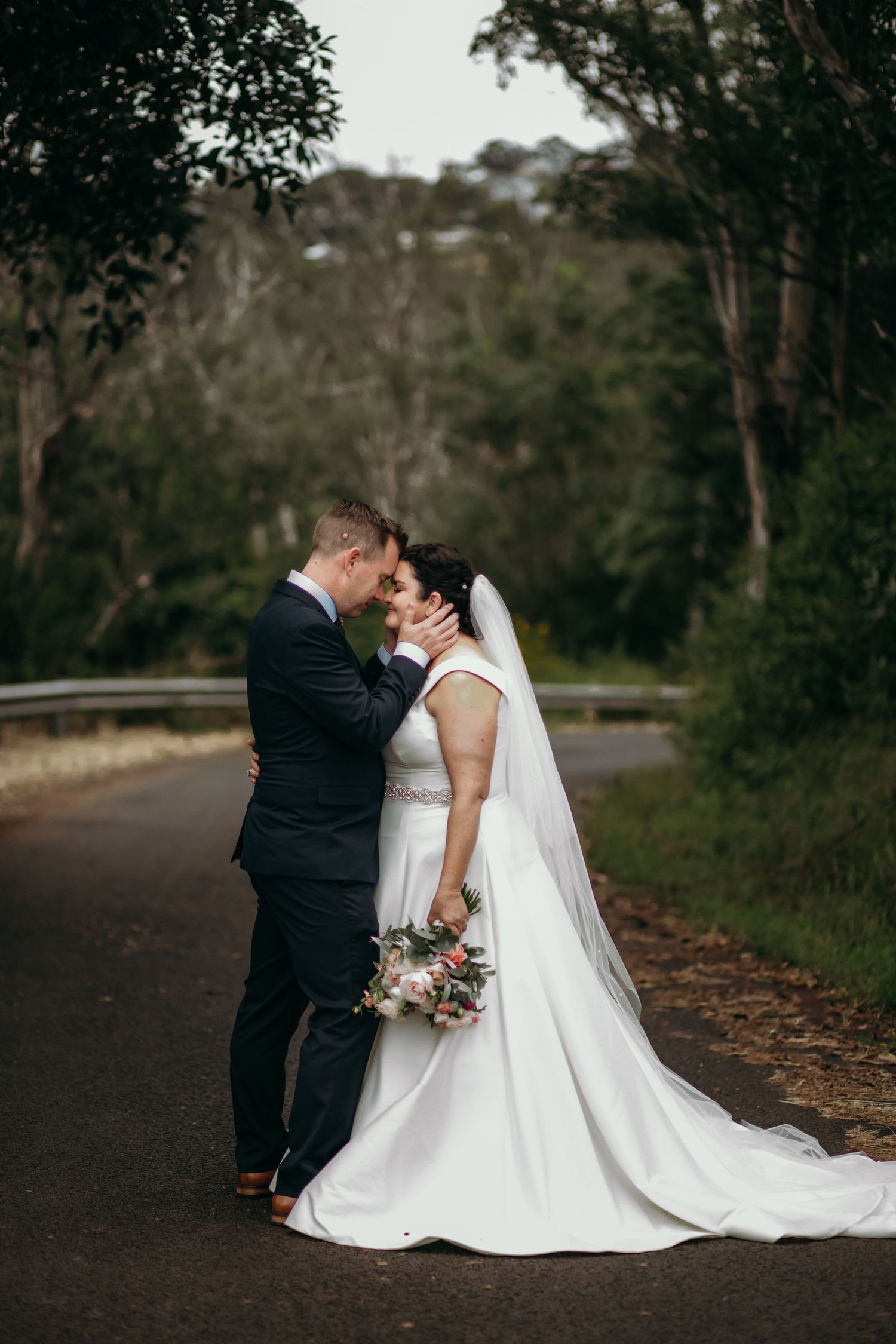 Couple in wedding attire standing together on a rustic path surrounded by trees.