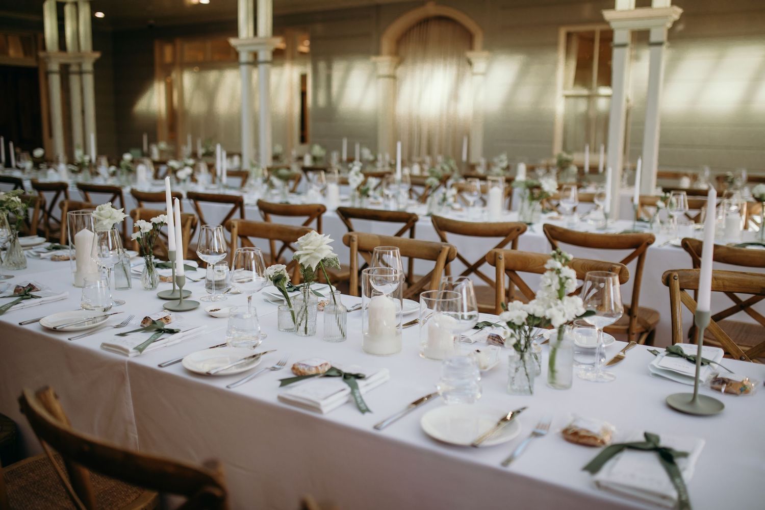 Elegant wedding reception tables with white linens, cross-back chairs, and delicate place settings in a bright banquet room.