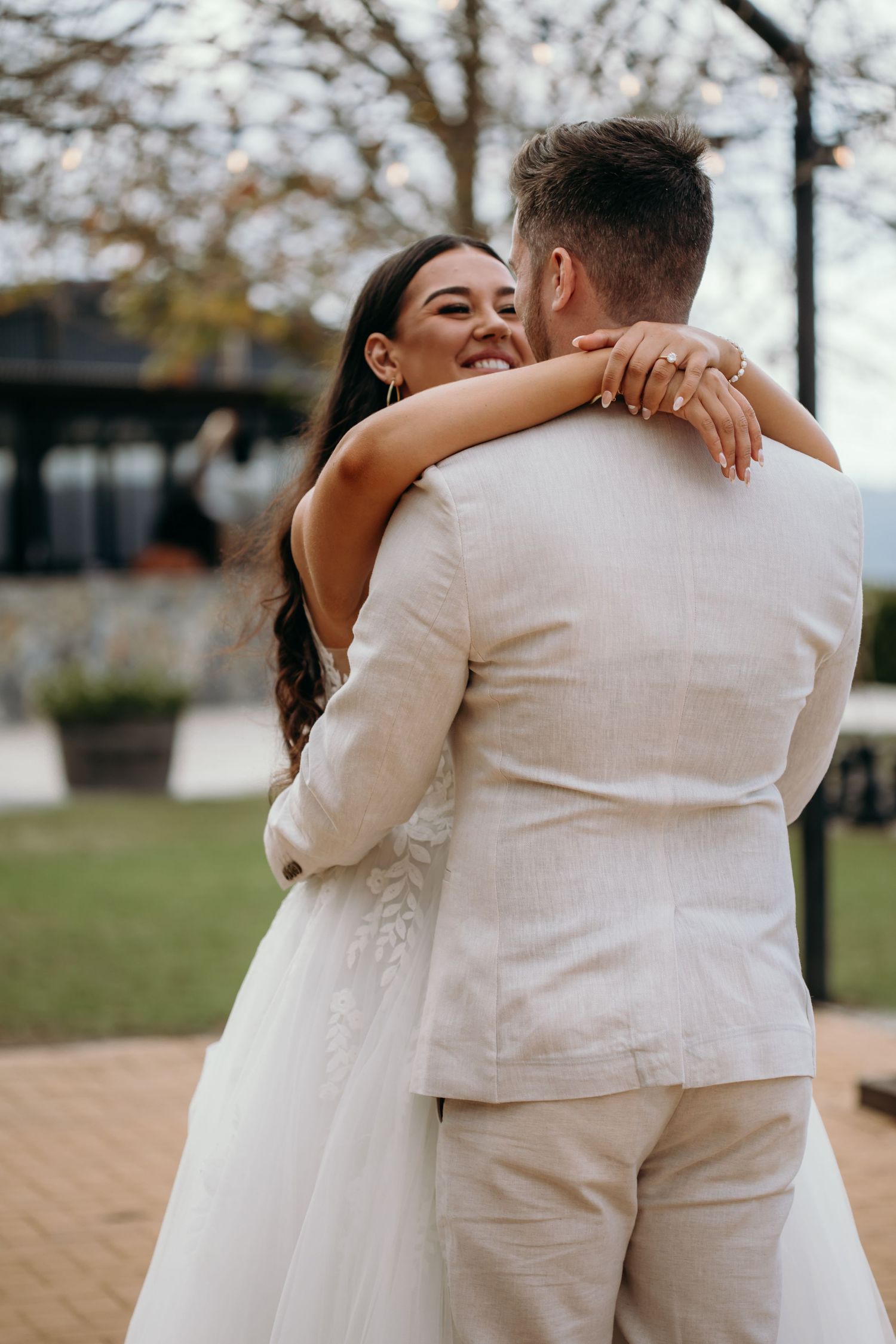 Newlywed couple embraces outdoors during their wedding day celebration with autumn trees in background.