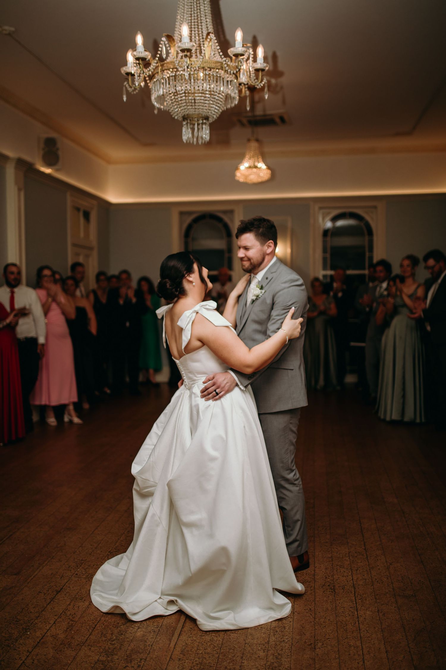 A couple shares their first dance at a wedding reception under elegant crystal chandeliers on a wooden dance floor.