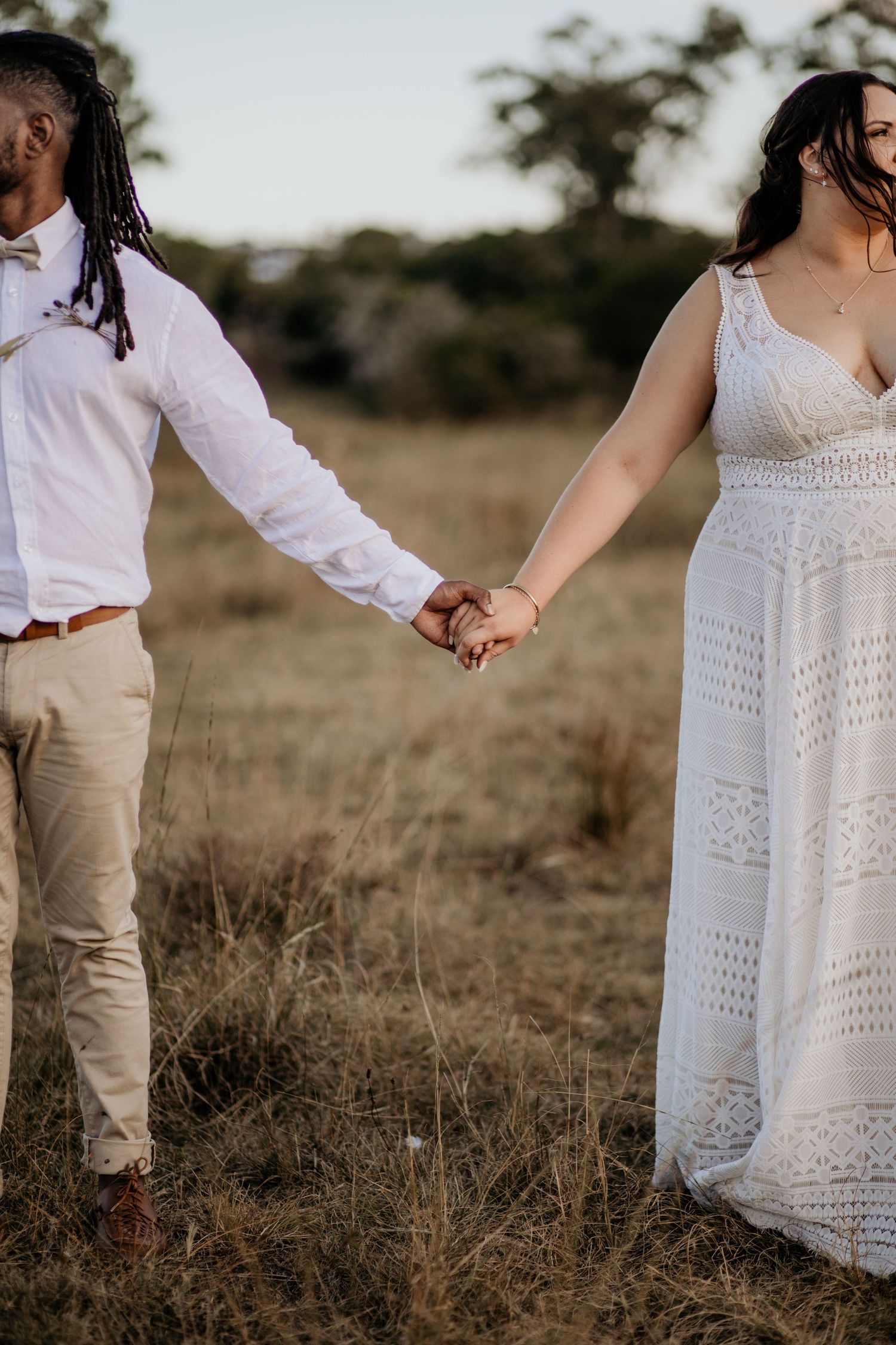Couple holding hands in field wearing casual white wedding attire.