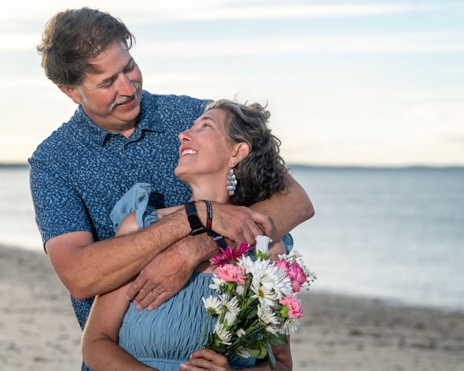 Couple embracing on beach, woman holding pink and white flowers, smiling joyfully at sunset.