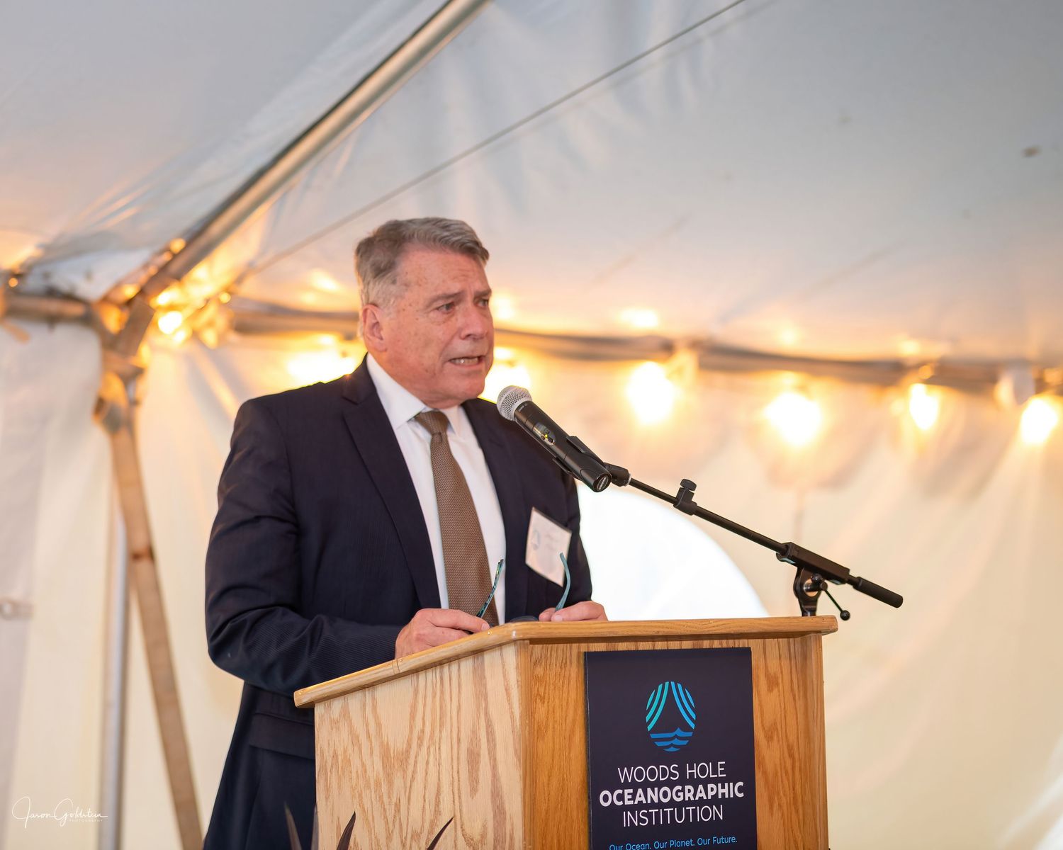 Speaker at a podium giving a presentation under an illuminated tent at a formal event.