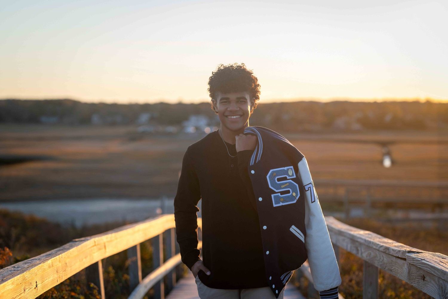 Young man in varsity jacket smiling at golden hour sunset overlooking scenic marshland.
