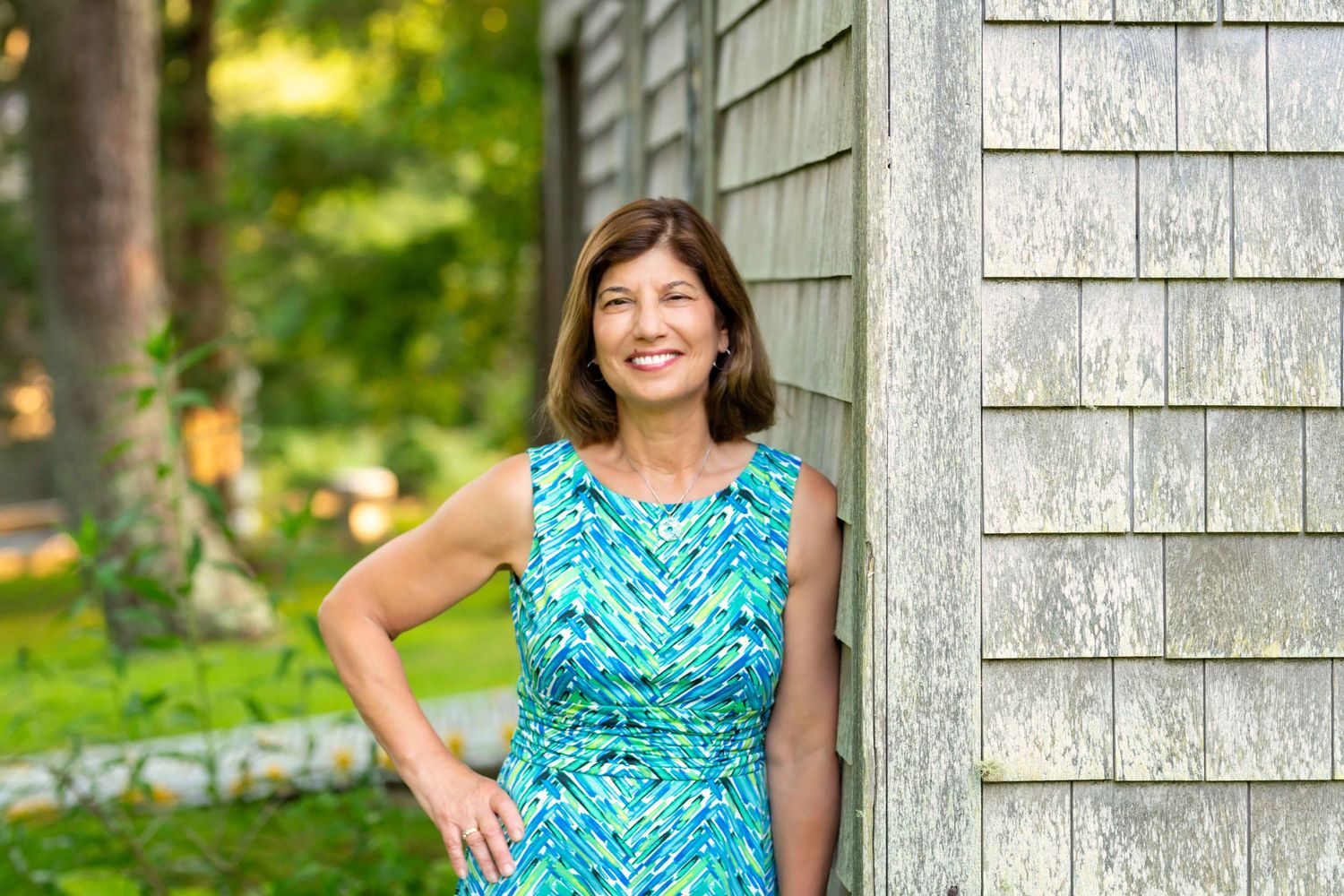 Smiling woman in blue floral dress leaning against weathered shingle wall in lush green garden.