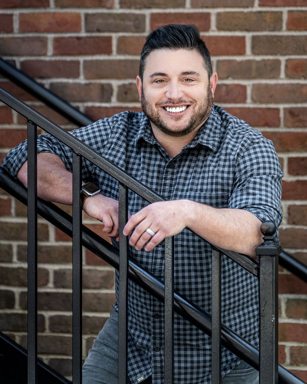 A person in a checkered shirt smiles while leaning on black metal railings against a brick wall background.
