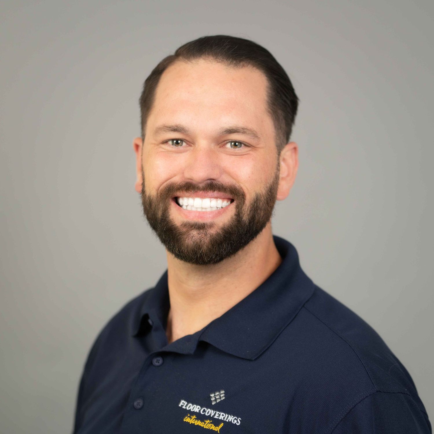 Smiling man with beard wearing a Floor Coverings International navy polo shirt against a gray background.