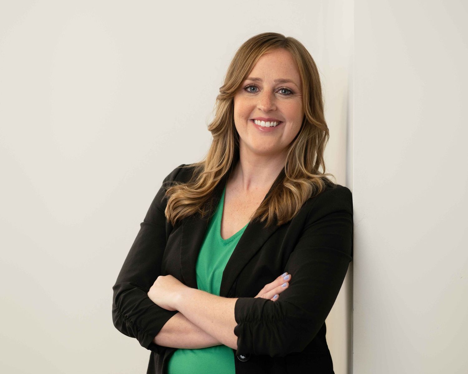 Smiling woman in black blazer and green top poses with arms crossed against a white background.