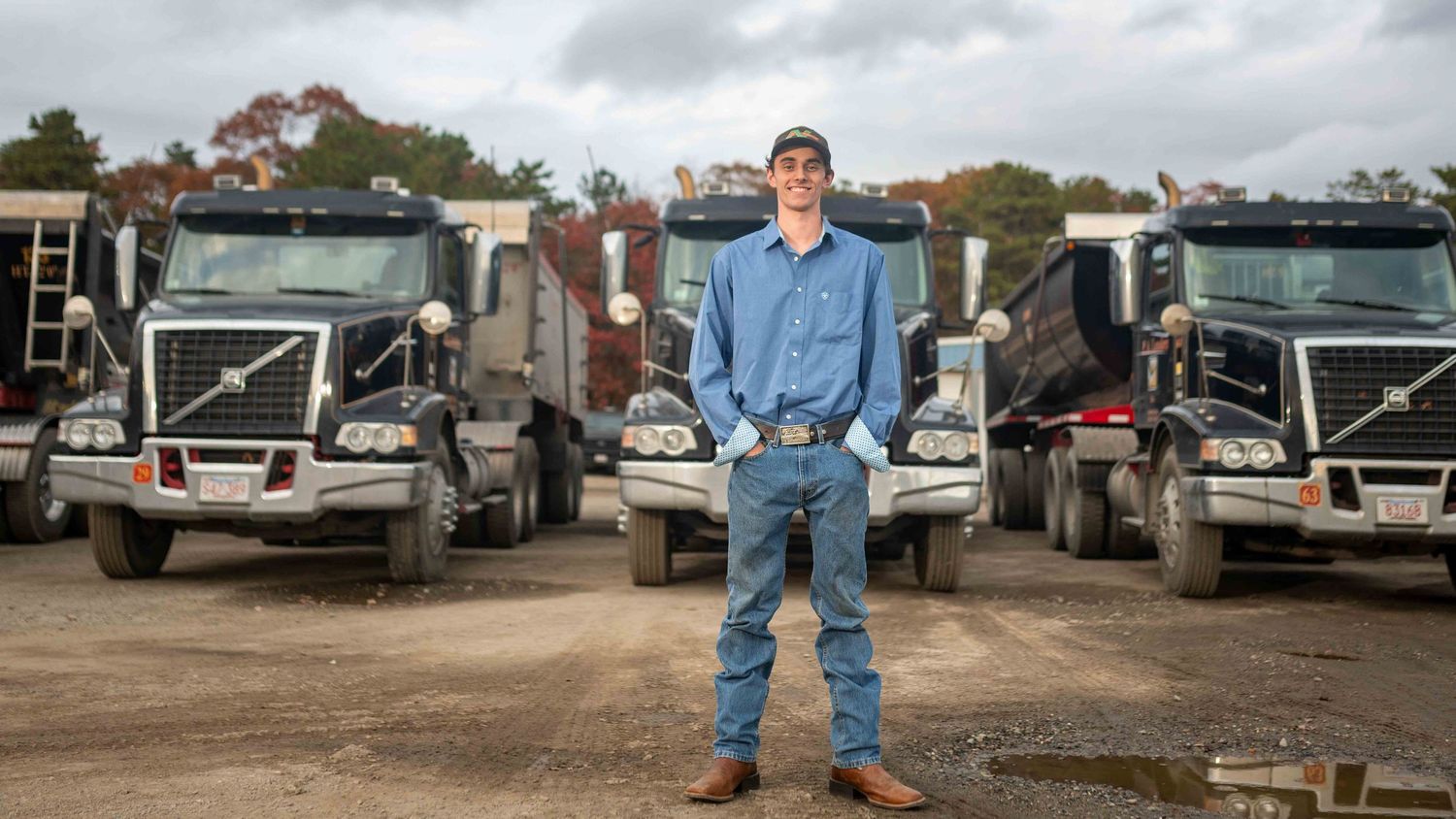 Man in blue shirt stands confidently before a fleet of large dump trucks in an outdoor yard.