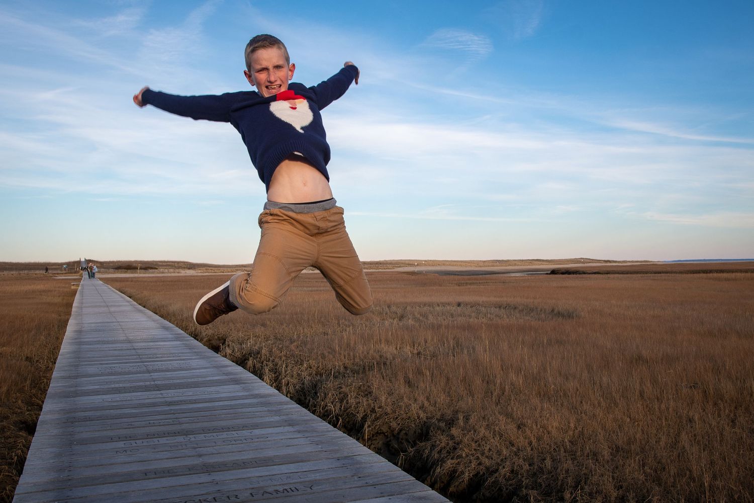 Wooden boardwalk stretches across a scenic marsh landscape under blue skies.