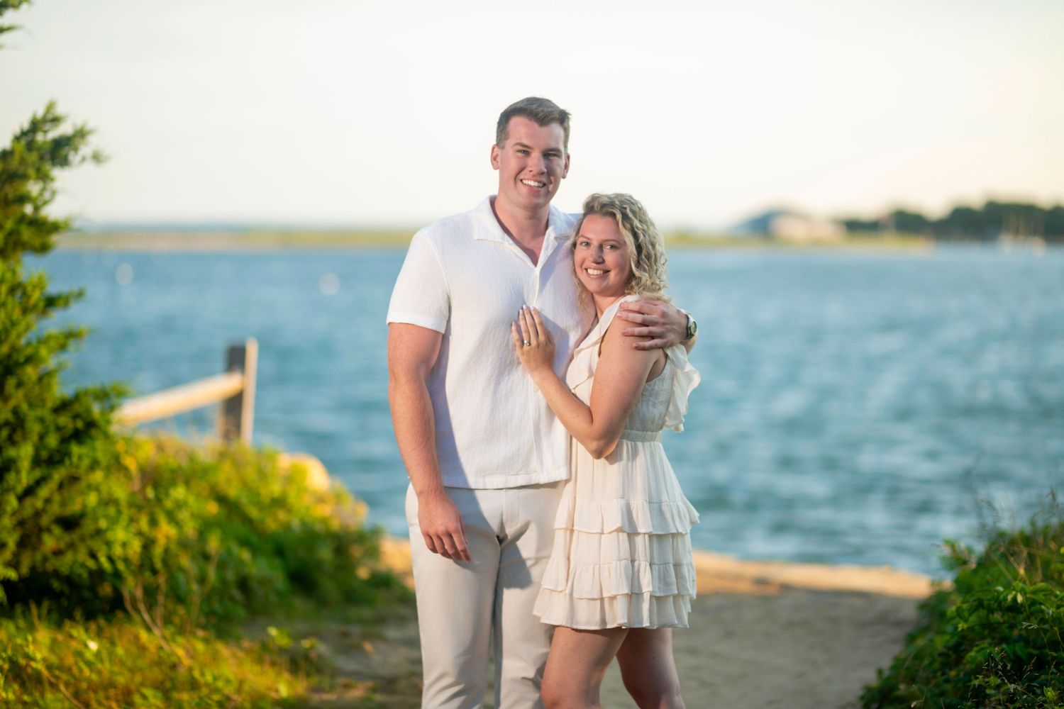 A couple shares a romantic moment on a beach at sunset with the ocean and coastline in the background.