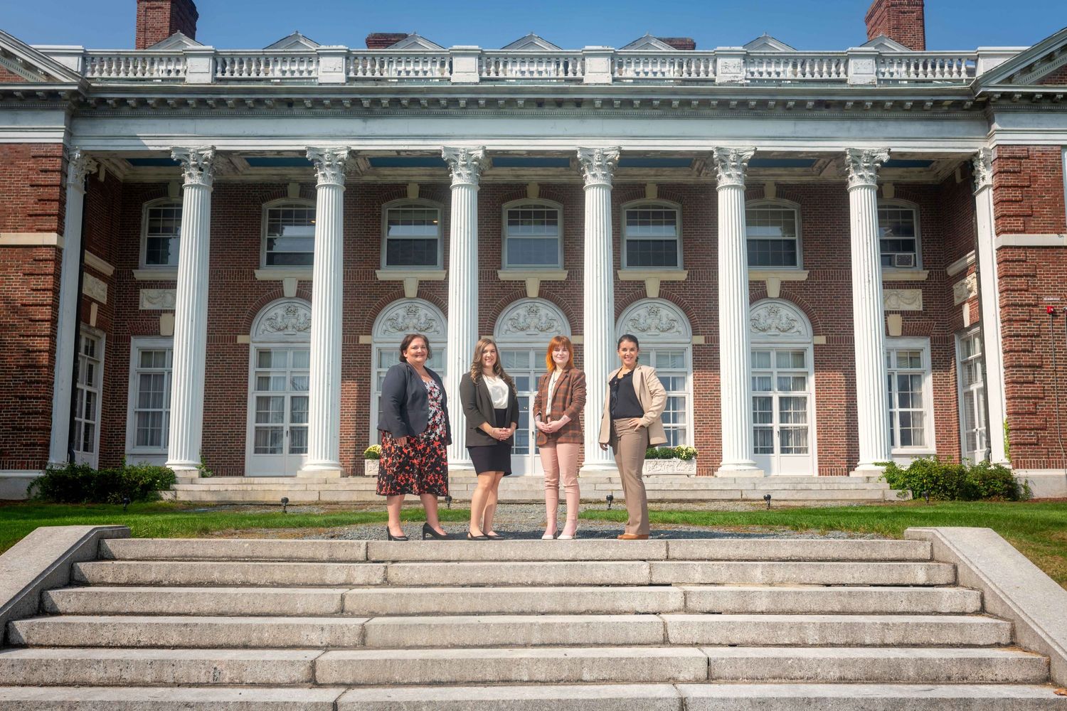 Four professionals pose on steps of a grand brick building with white columns on a sunny day.