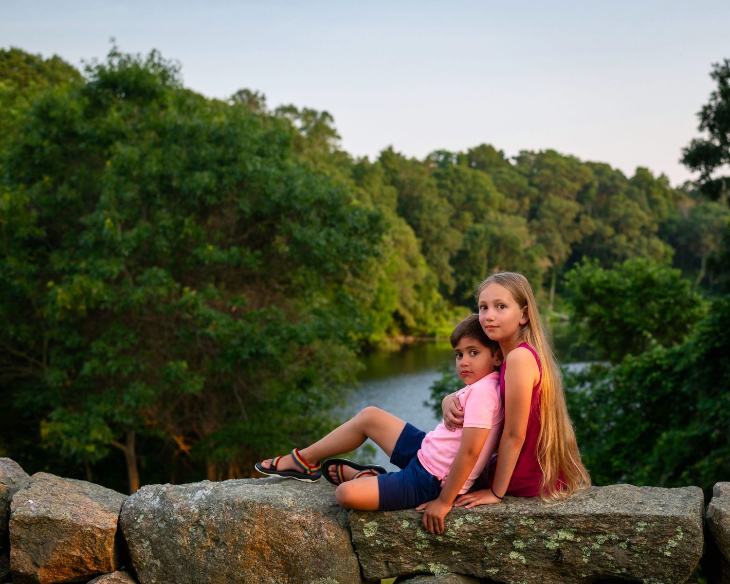 People relaxing on a rustic stone wall with lush green trees in the background during golden hour.