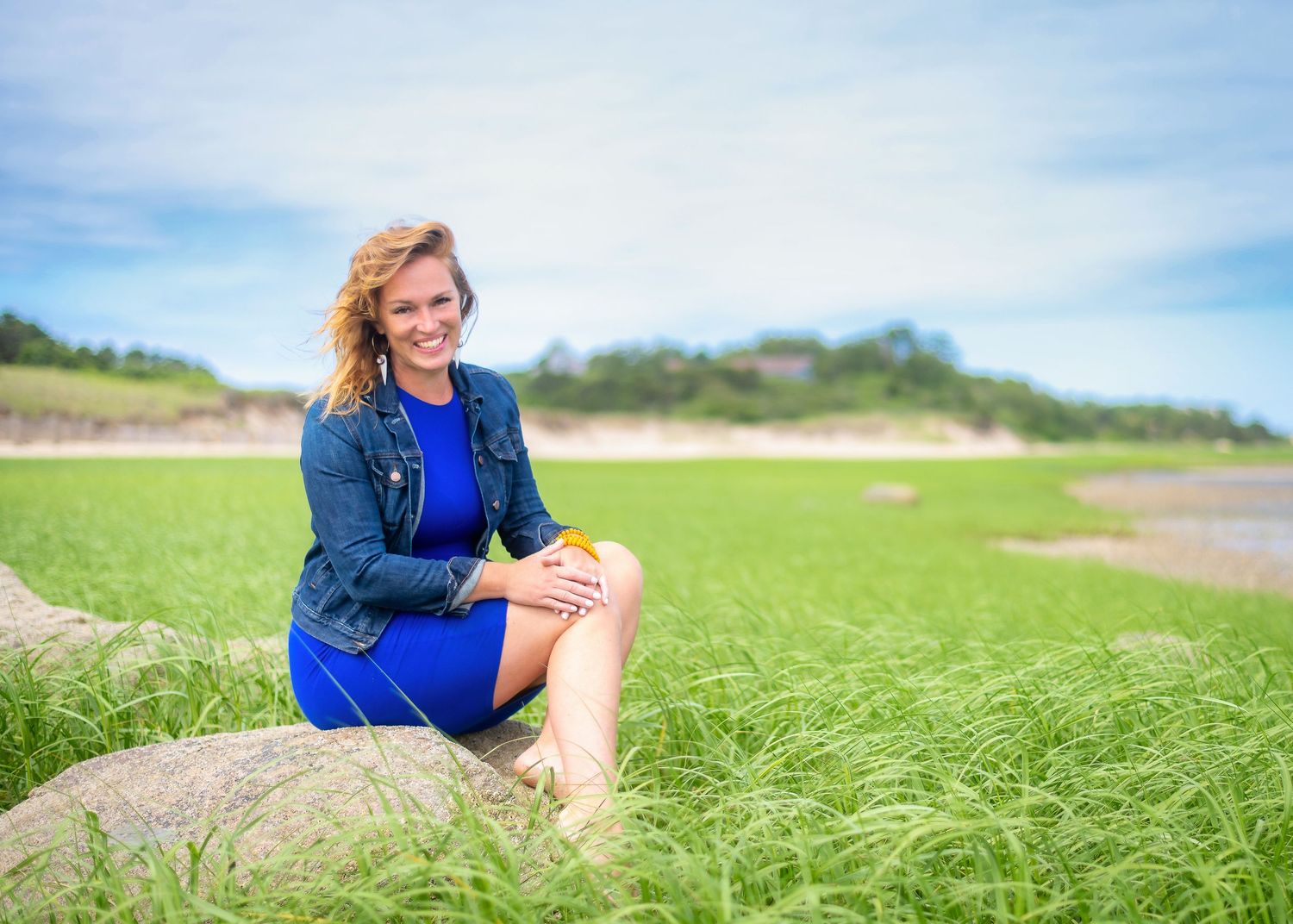 Woman in blue outfit posing in green field with coastal scenery in background.