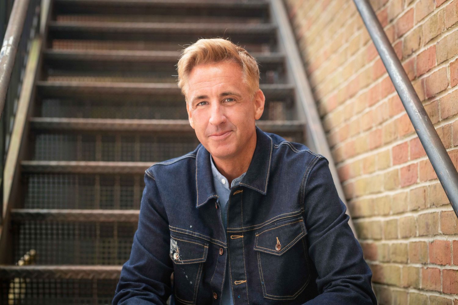 Smiling middle-aged man in a denim jacket sitting on stairs against a brick wall background.