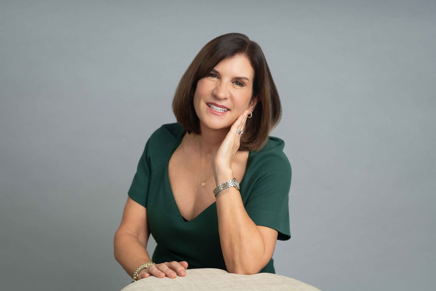 Woman in green dress poses elegantly with hand on cheek, smiling confidently against a gray studio background.