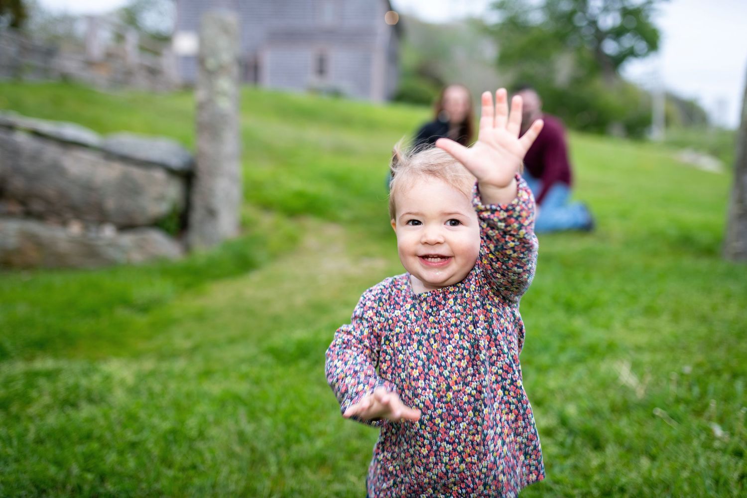 A toddler in a leopard print dress plays and waves while sitting on grassy hill in a park setting.