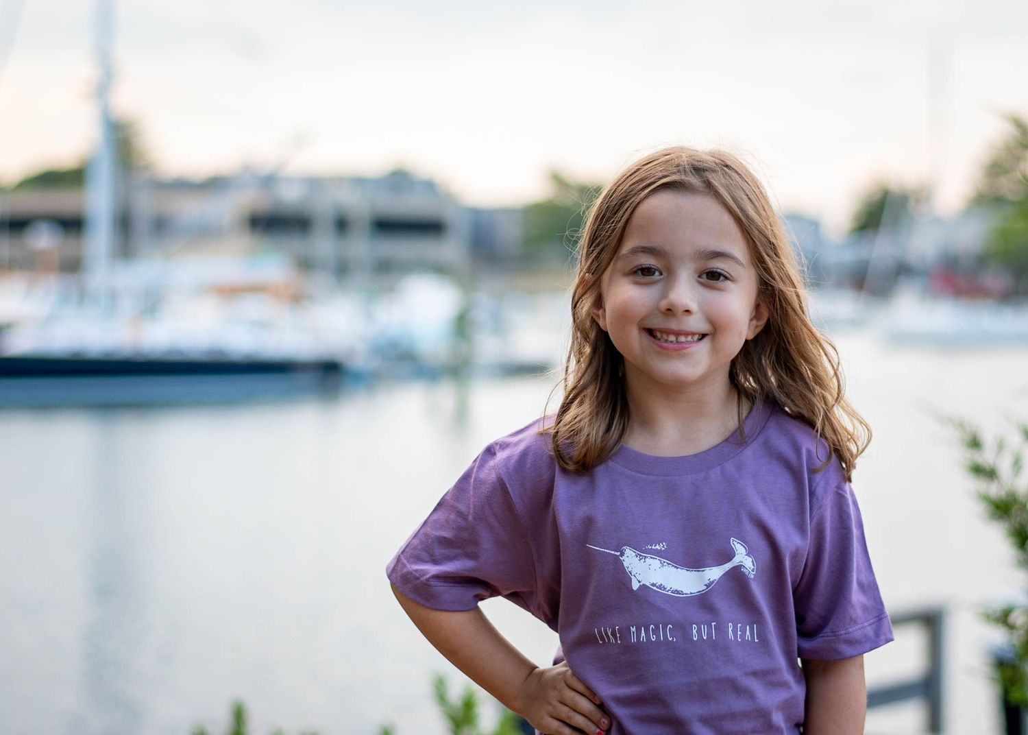 Young person in purple t-shirt smiling at marina with boats in background during golden hour.