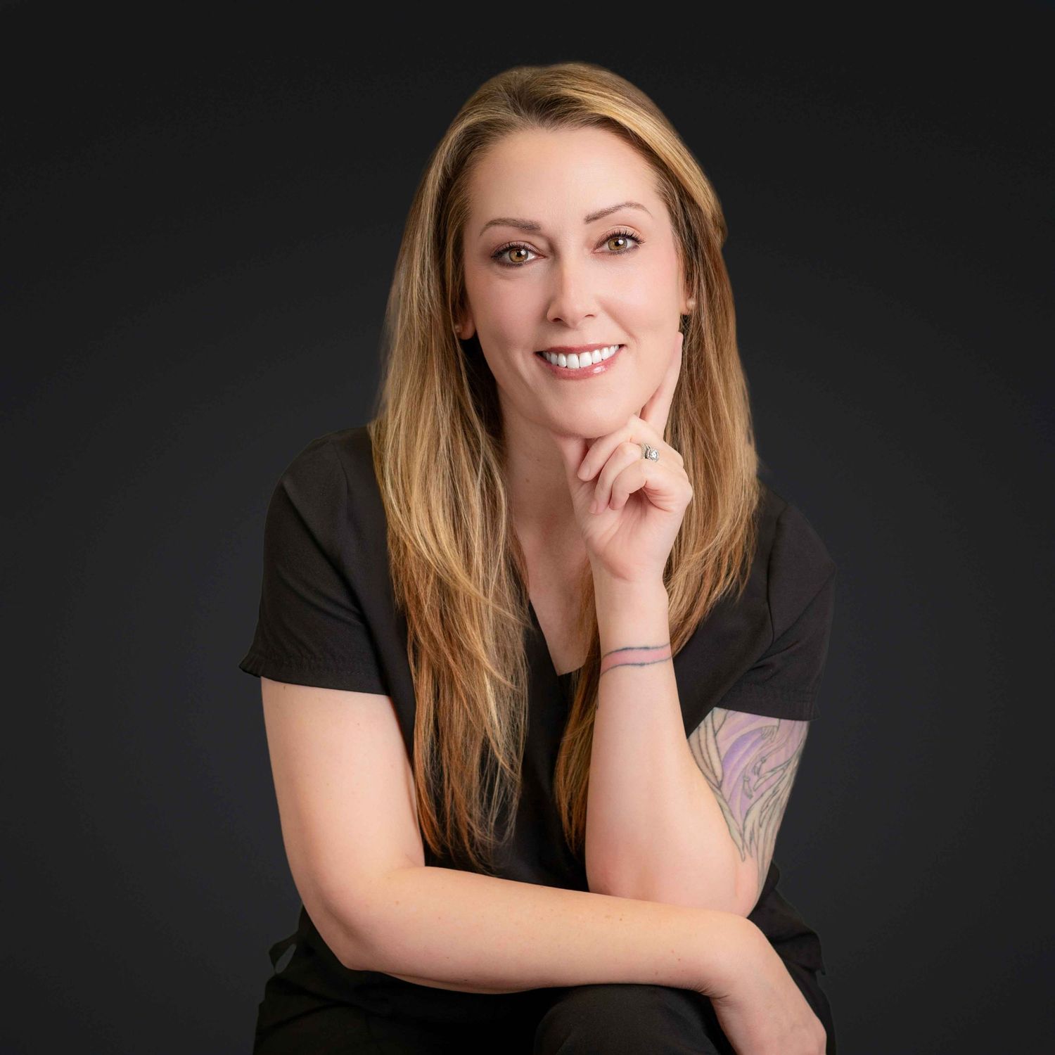 Smiling blonde woman in black top posing confidently with hand on chin against dark background.