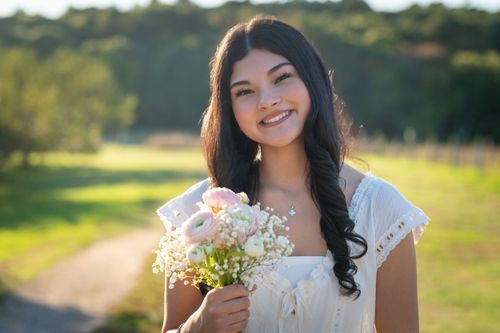 Someone in a white lace dress holds a delicate bouquet of pink and white flowers in a sunny outdoor setting.