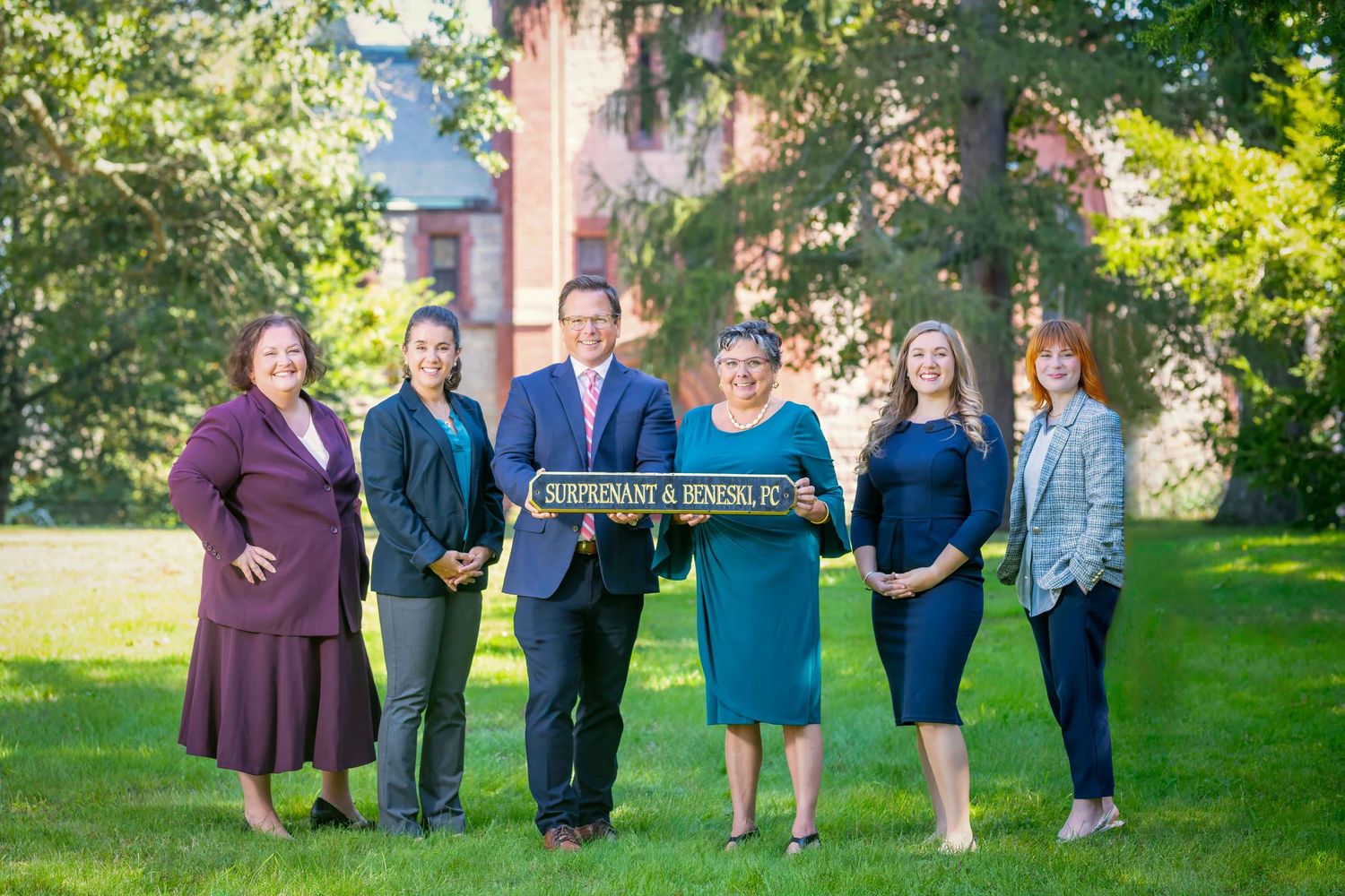 Professional law firm team of six smiling colleagues posing outdoors on green lawn holding office sign.