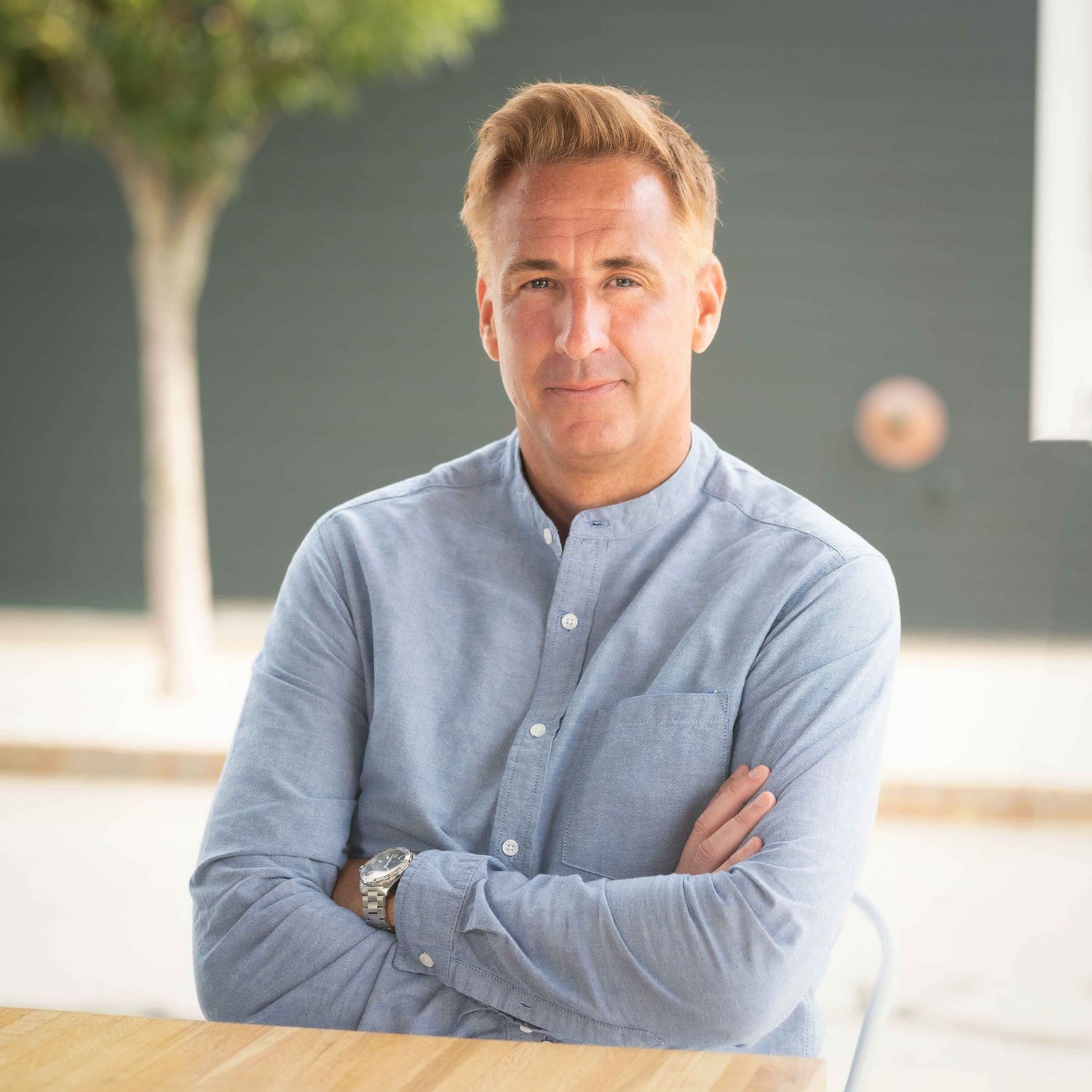 Man in light blue shirt sitting with arms crossed, smiling confidently in a modern bright interior space.