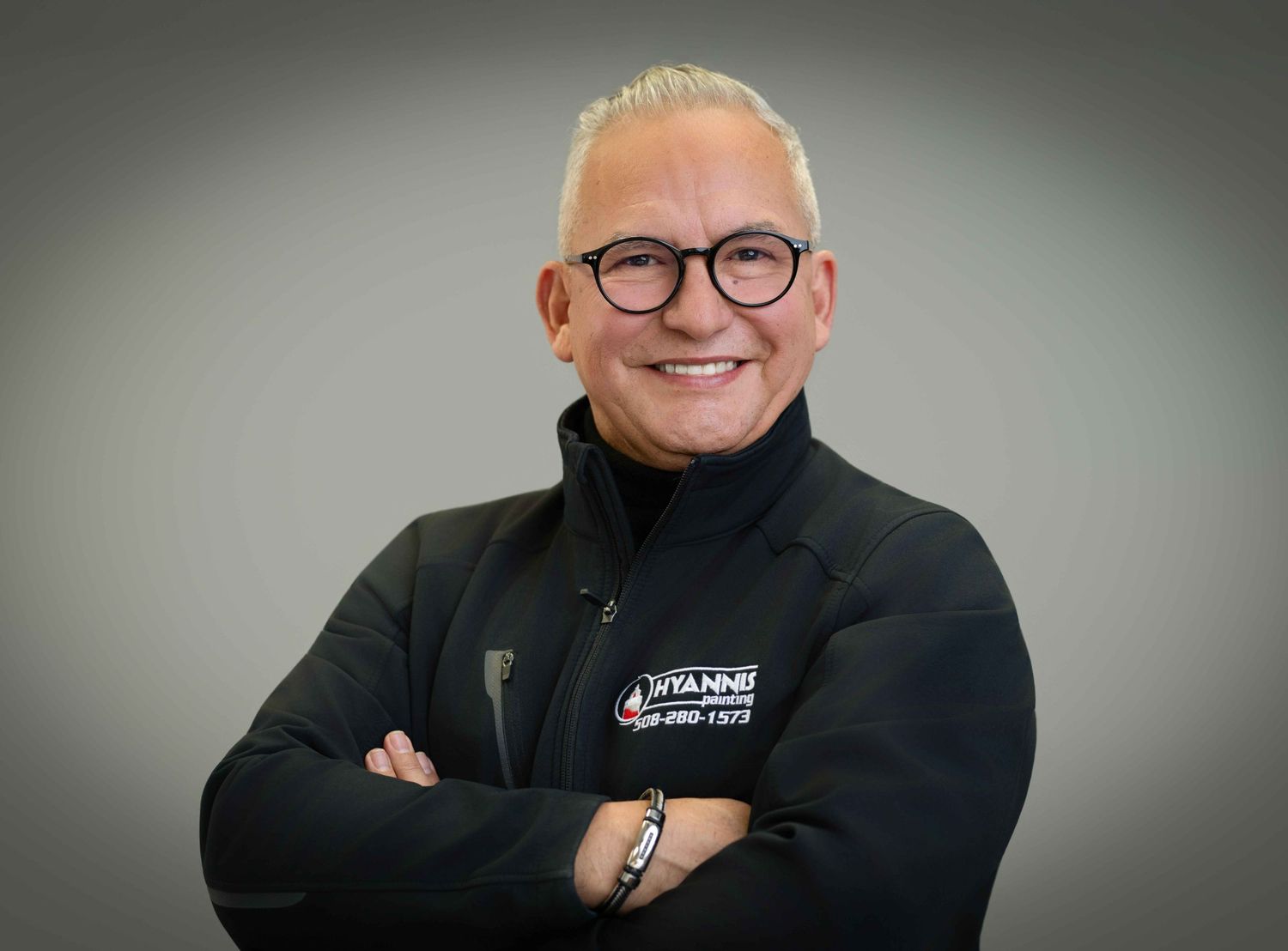 Smiling professional man with gray hair and glasses wearing a black company jacket with arms crossed.