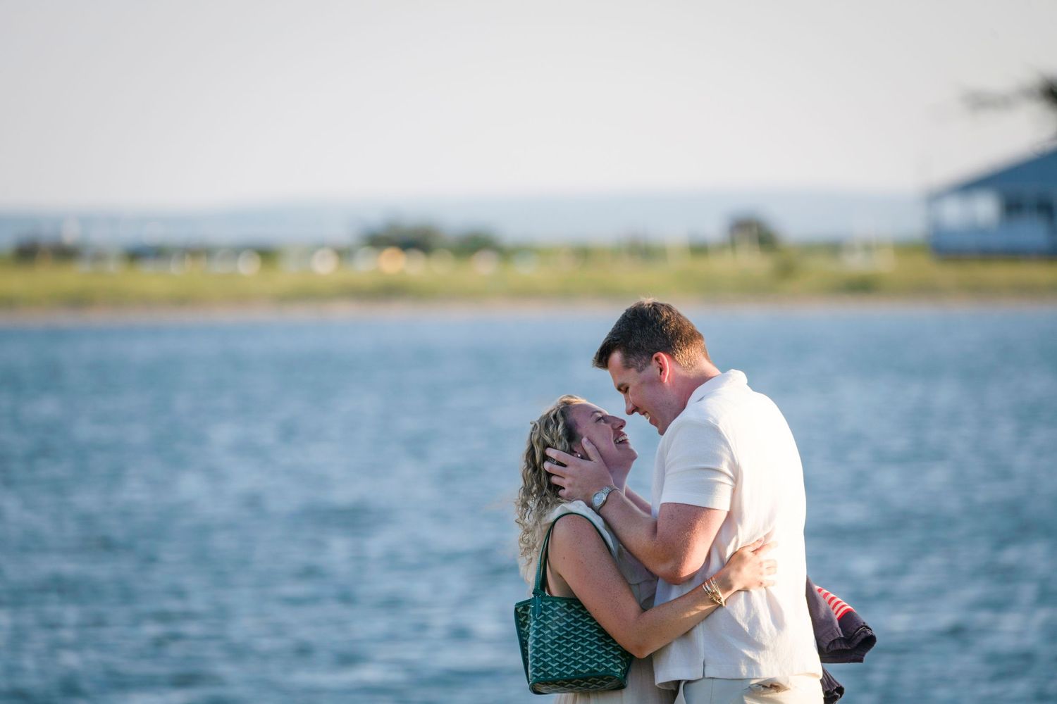 Two people embrace while standing on a scenic coastal path with boats and water in the distance.