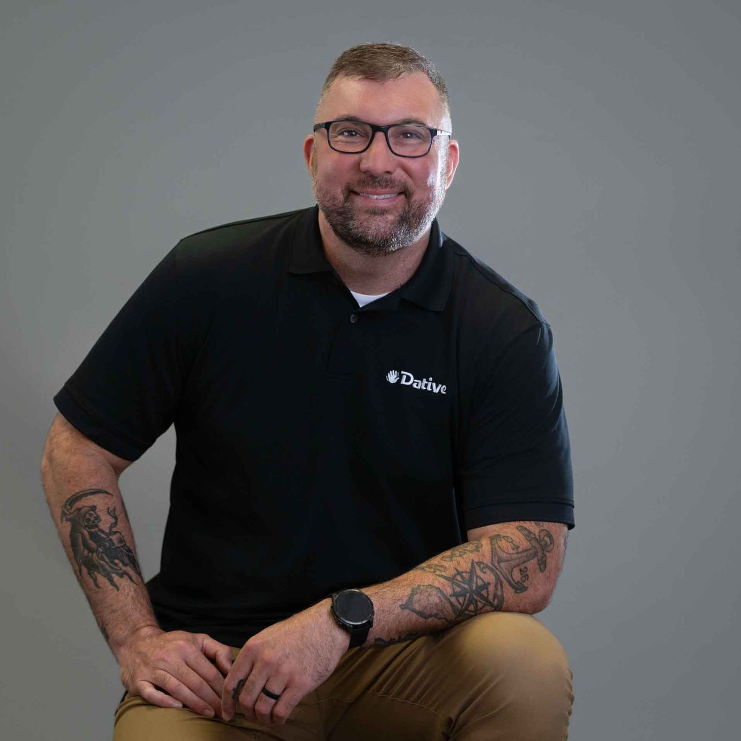 Man in black Datlan polo shirt with tattoos smiling, seated against gray background.
