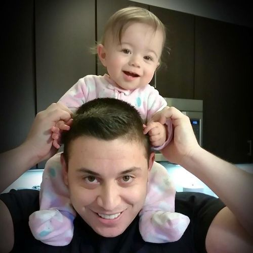 A smiling baby sits on top of someone's shoulders in a dimly lit indoor setting.