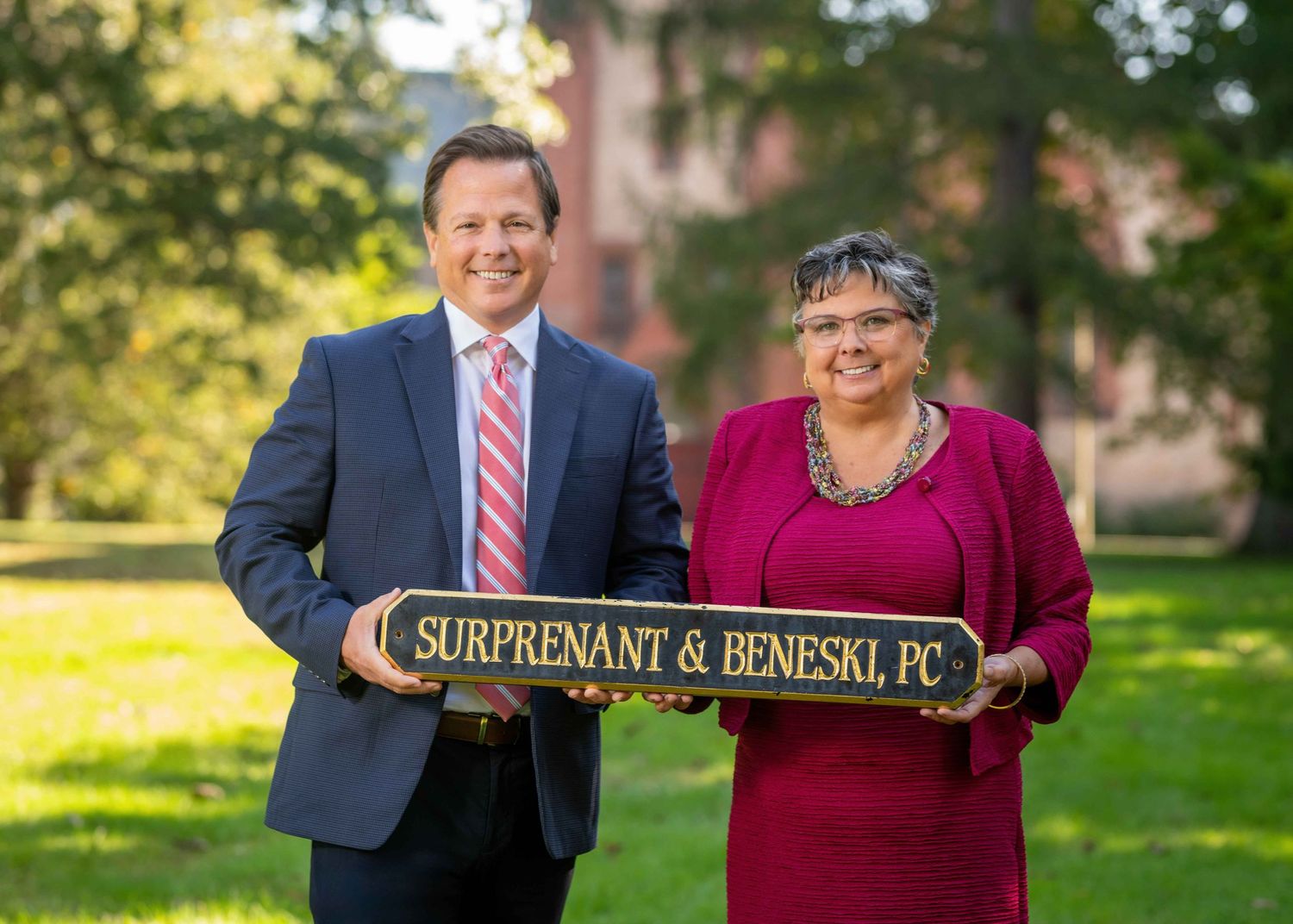 Two professionals holding a Surprenant & Beneski PC sign outdoors on a sunny day at a law firm.