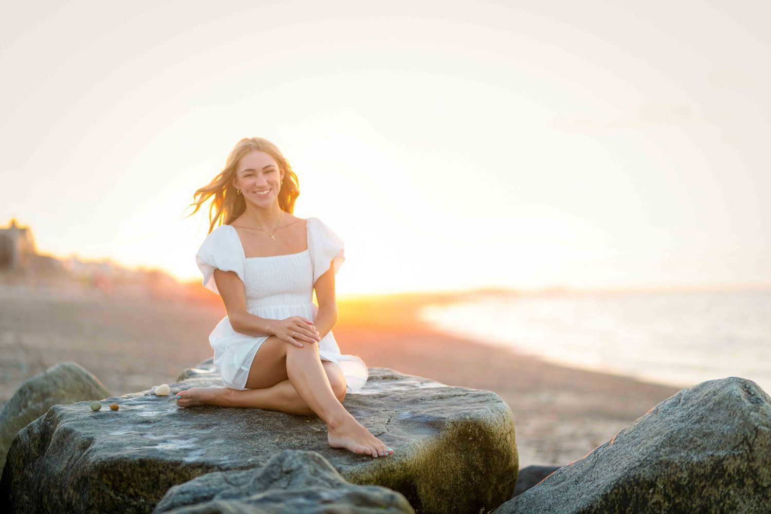 Young woman in white dress sitting on rocks at beach during golden sunset with waves in background.