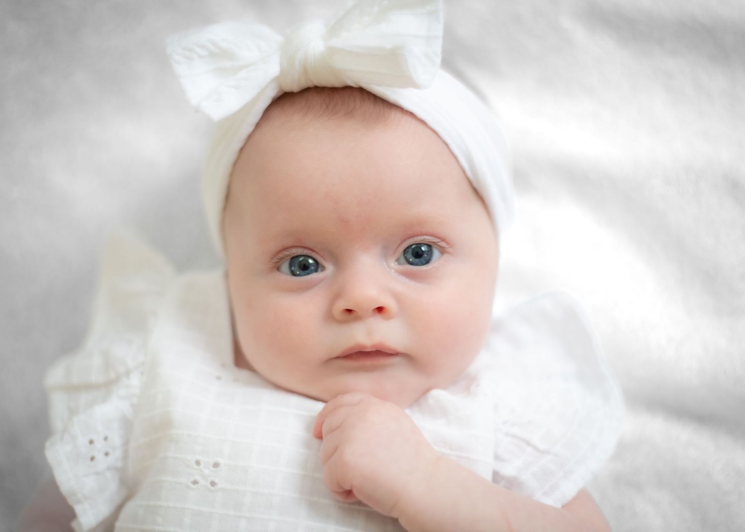 Baby wearing white bow headband and dress against soft white background.