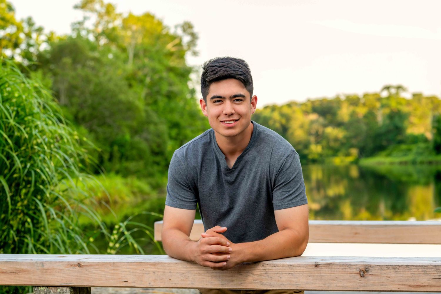 Young man in gray t-shirt leaning on wooden railing with lush green trees and serene lake in background.