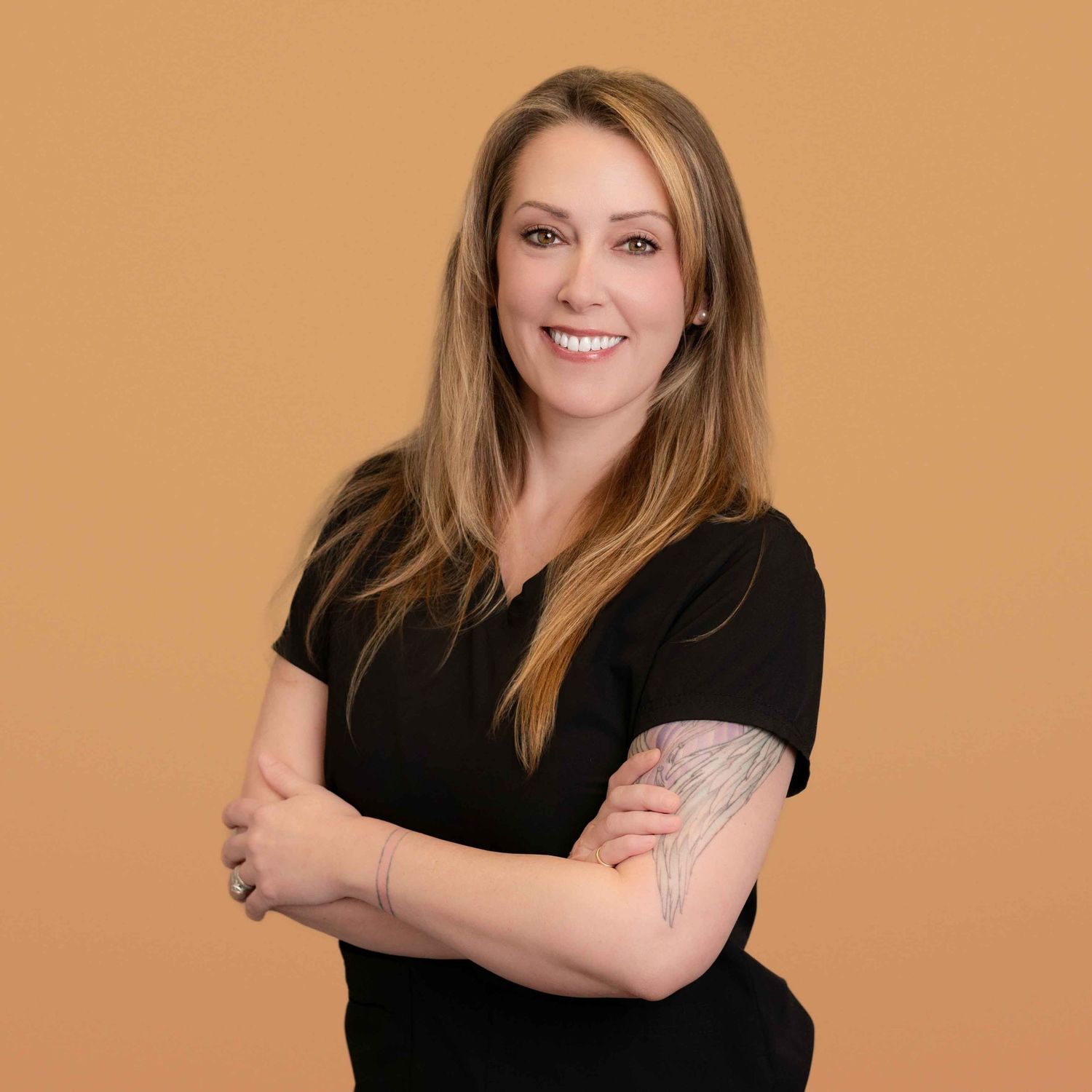 Smiling woman with long blonde hair, arms crossed, wearing a black top, with a tattoo on her arm, against an orange background.