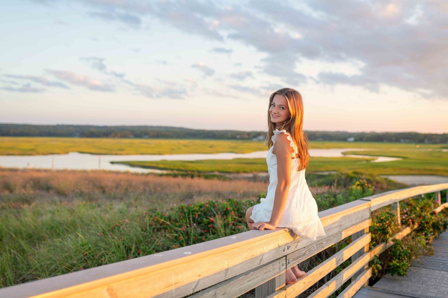 Young girl in white dress smiling on wooden boardwalk overlooking scenic marsh at golden sunset.
