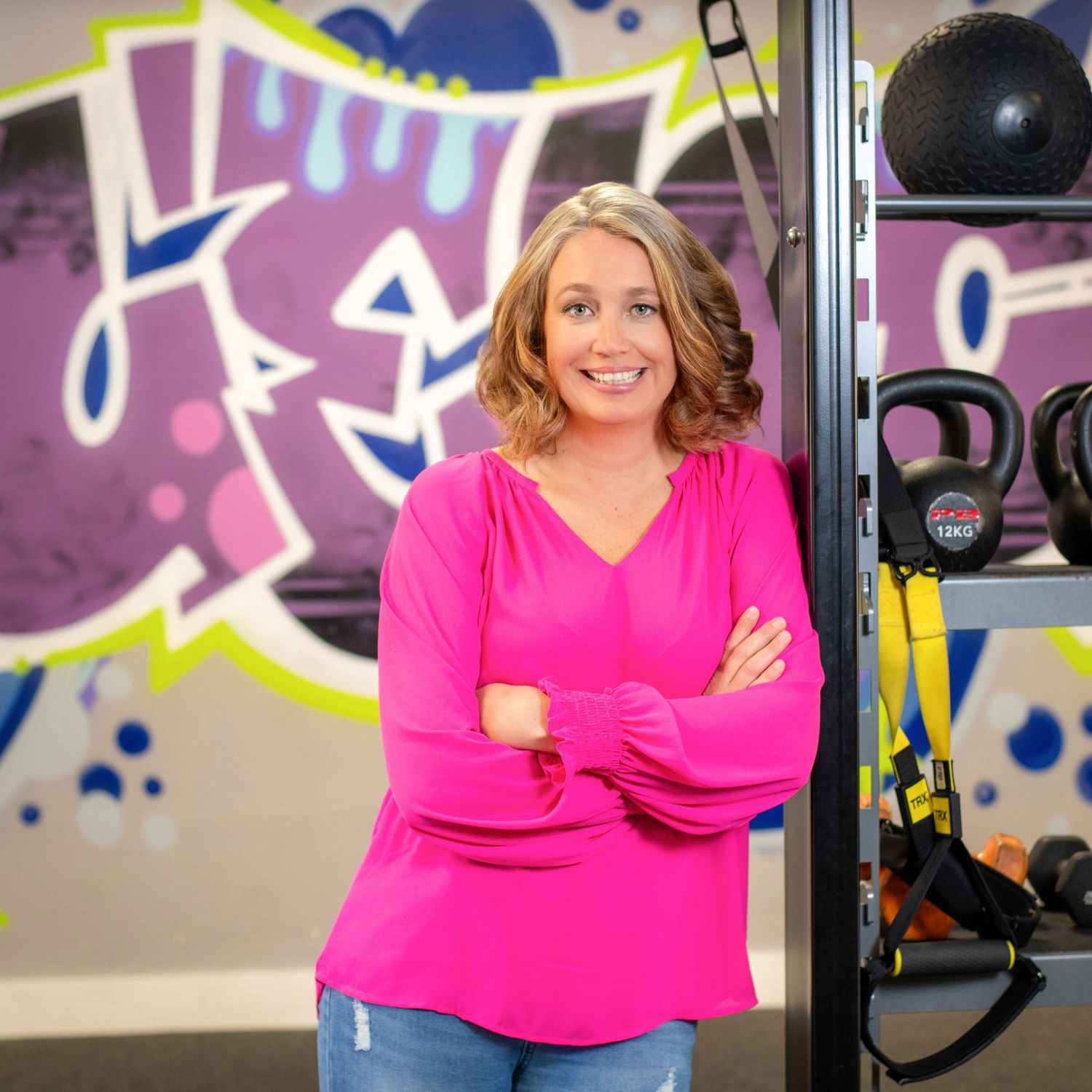 Woman in pink top poses confidently in a colorful gym with kettlebells and TRX equipment in the background.
