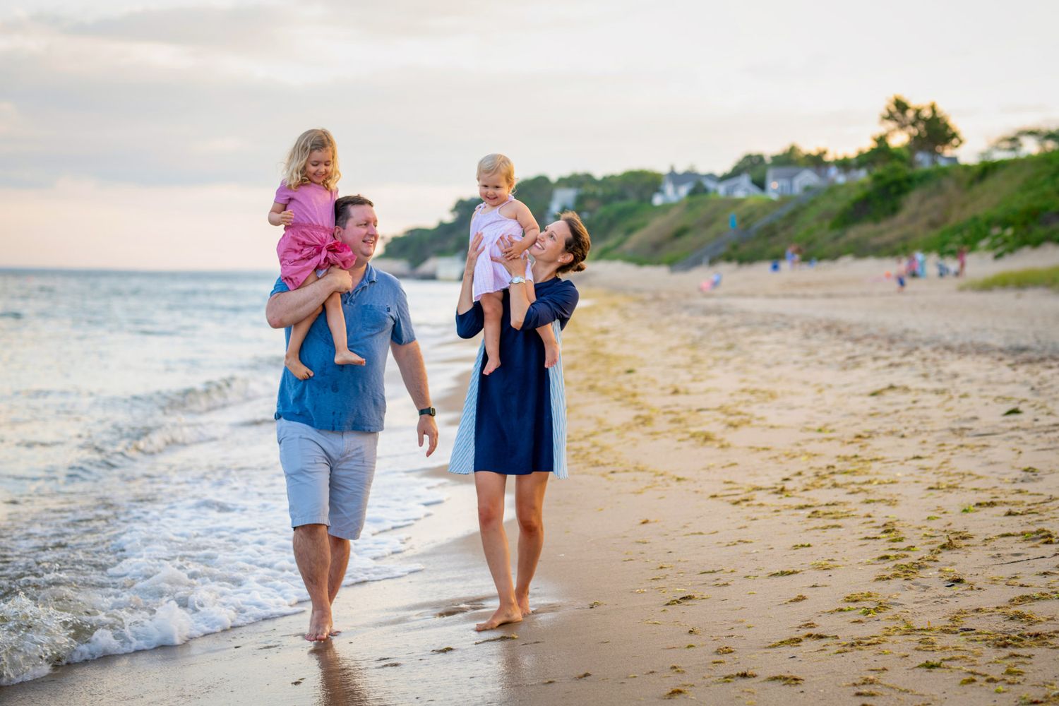 Family of four walking on a sandy beach at sunset, parents carrying young children on their shoulders.