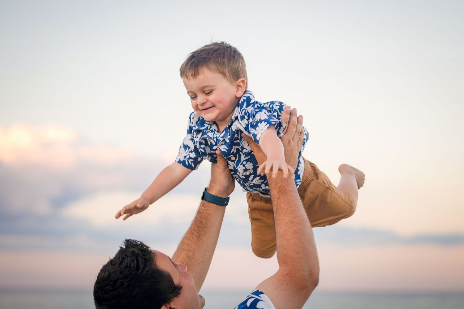 Parent lifting and playing with baby against sunset sky at beach.