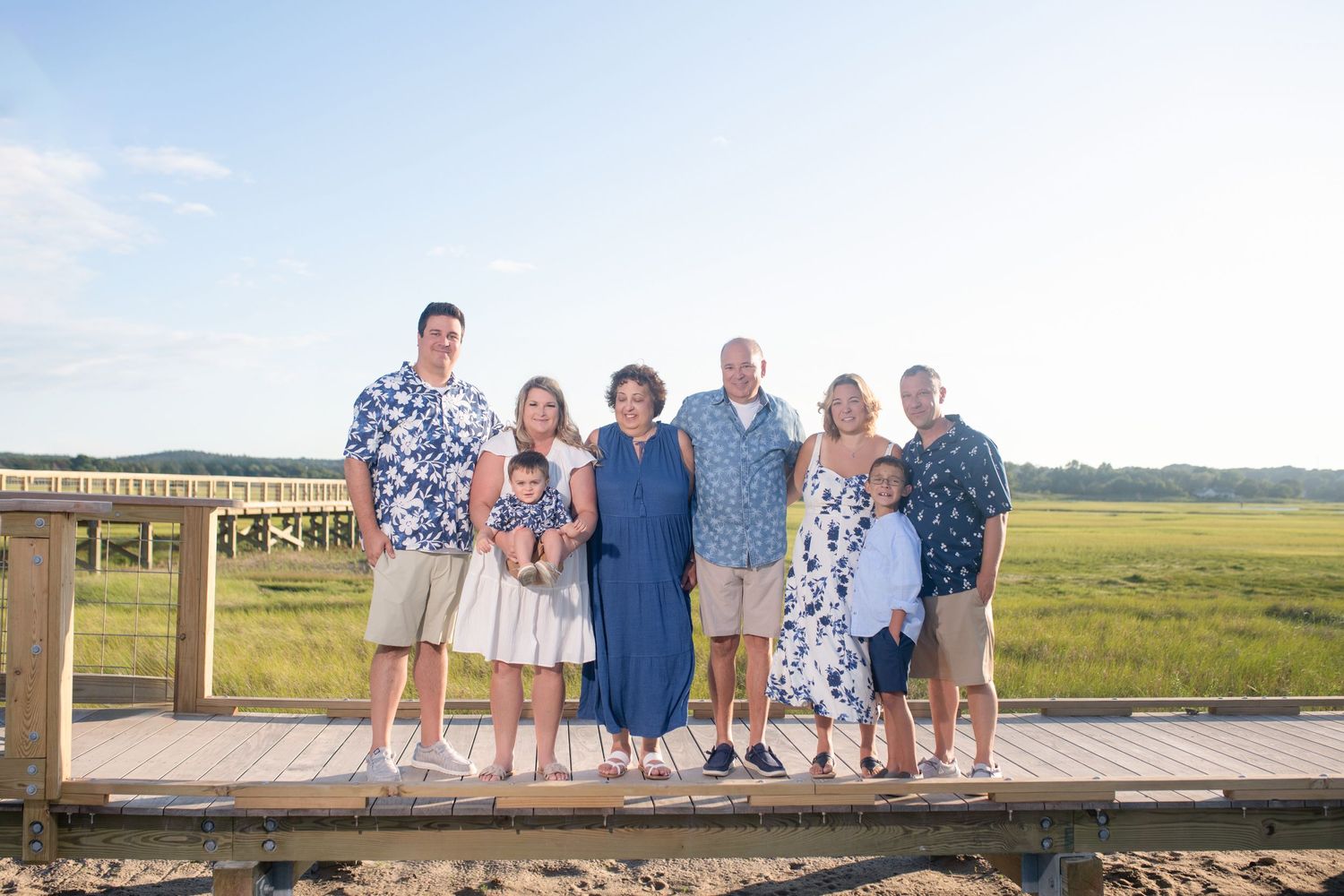 Extended family in coordinated blue and white outfits posing on wooden boardwalk near marsh.