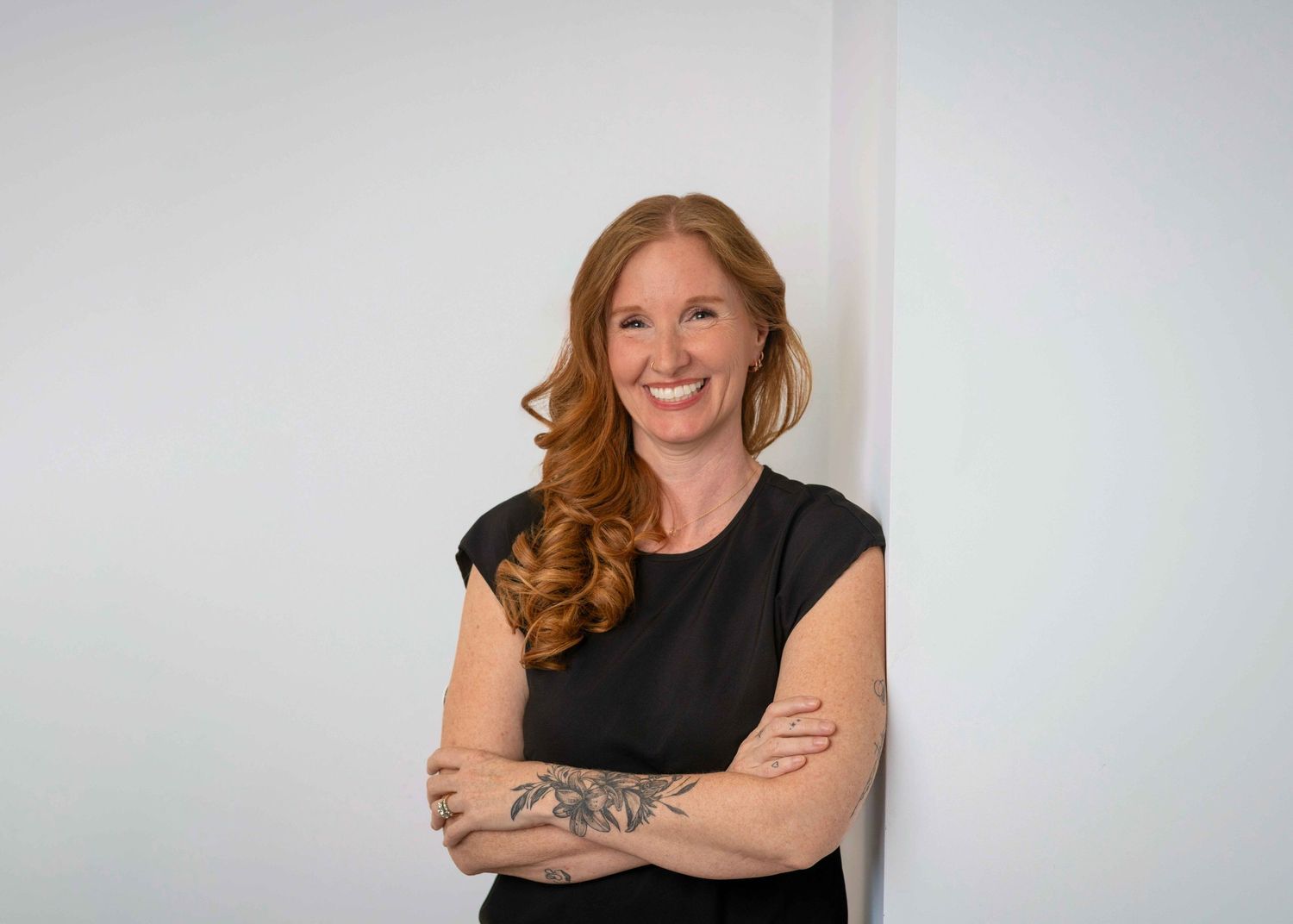 Smiling redhead woman with tattoos in black top leaning against white wall.