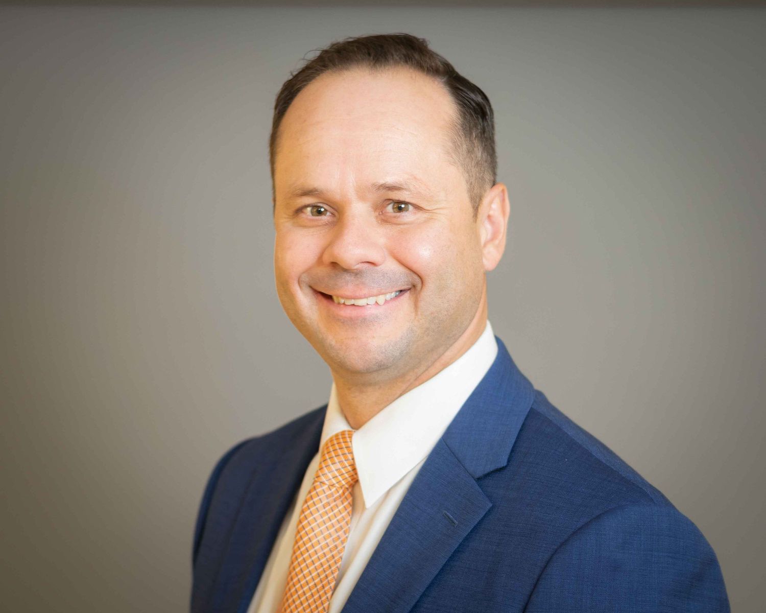 Professional man in blue suit and orange tie smiling confidently against a gray studio background.