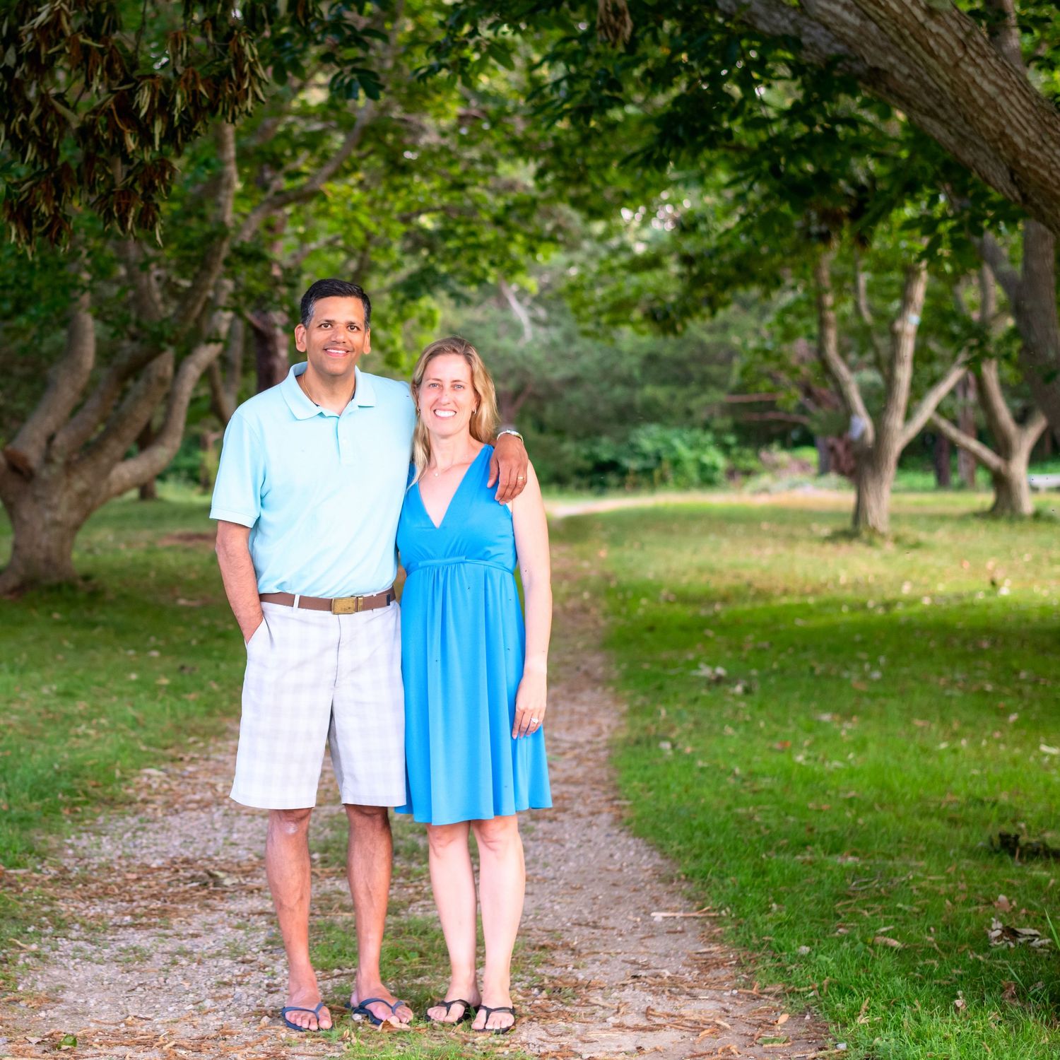 Family walking together on tree-lined dirt path in park setting.