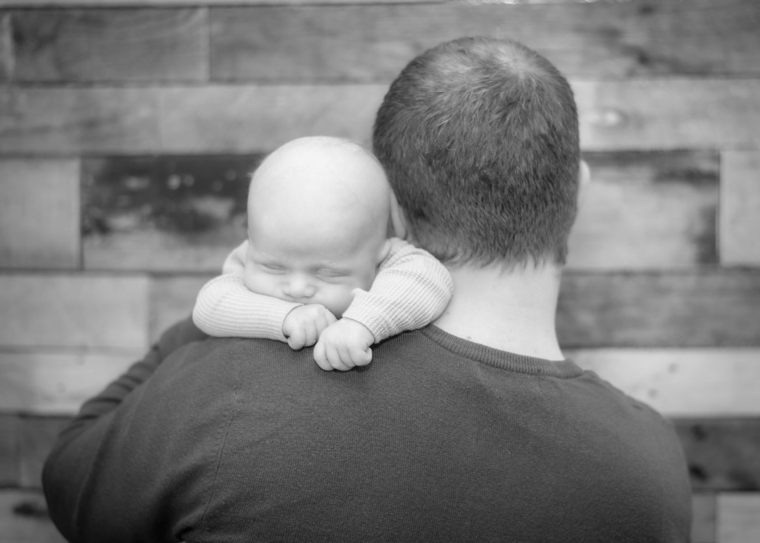 Black and white image showing baby resting on shoulder against wooden wall.
