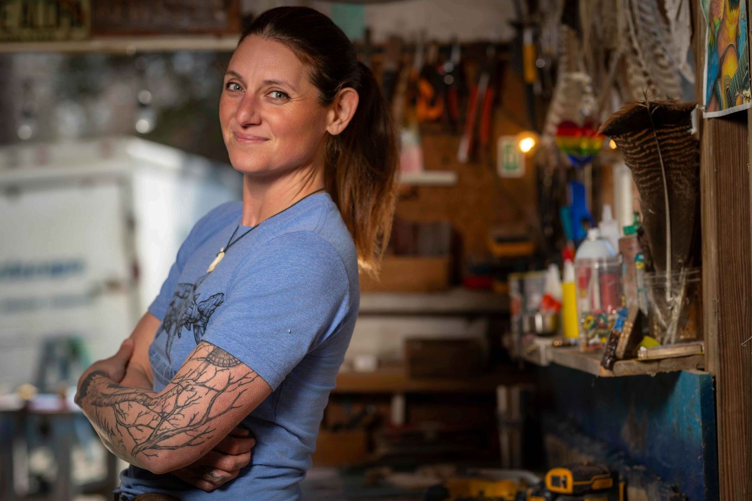 Tattooed woman with arms crossed standing confidently in a workshop surrounded by tools.