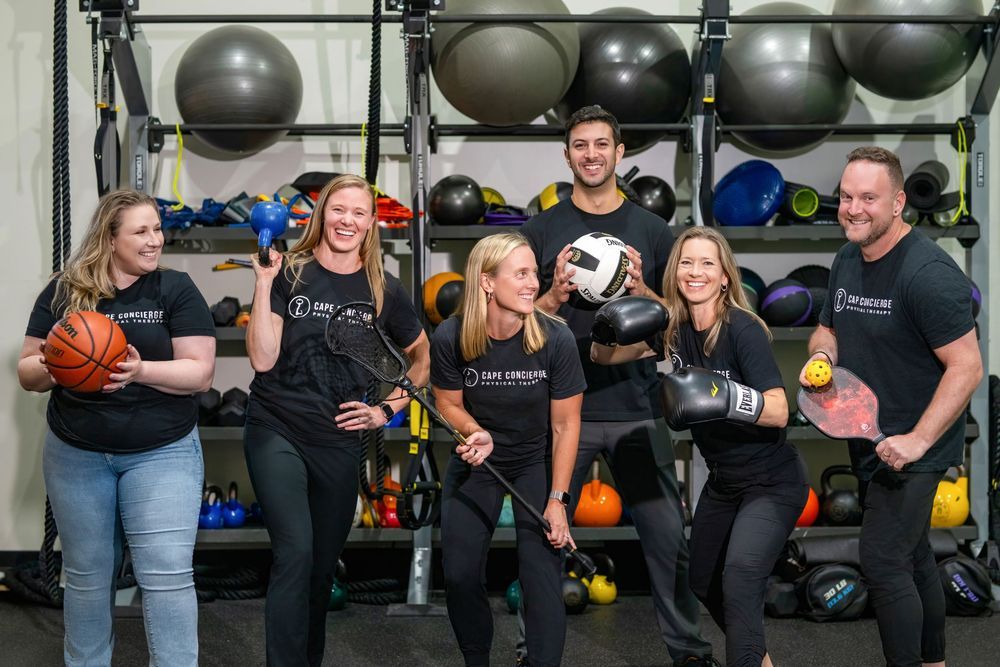Six fitness professionals pose with sports equipment in a gym, smiling in matching black branded workout shirts.